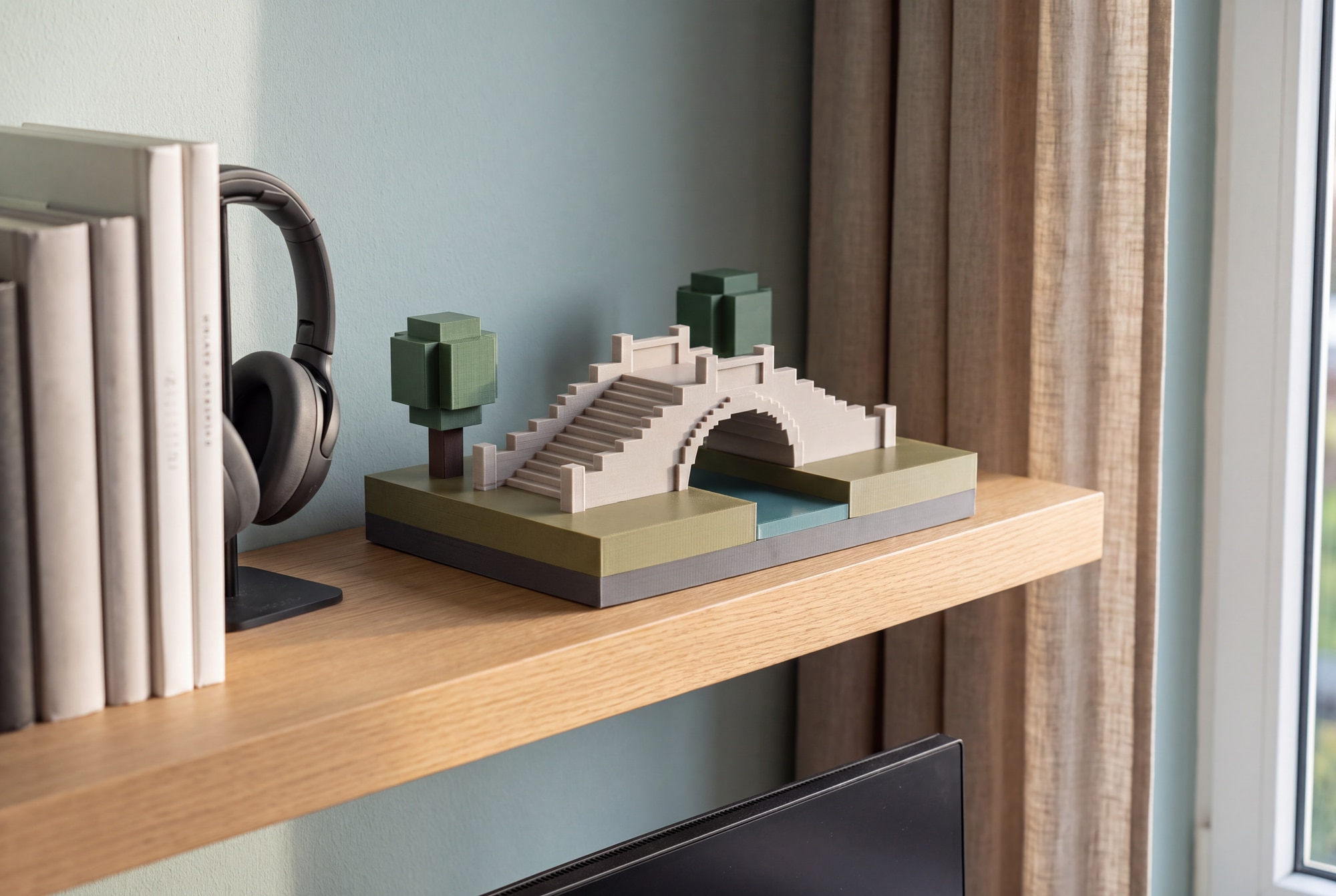 Illustration d’une maquette de pont à arches, avec deux arbres et son terrain entier, sur une étagère près de livres et d’un casque audio.